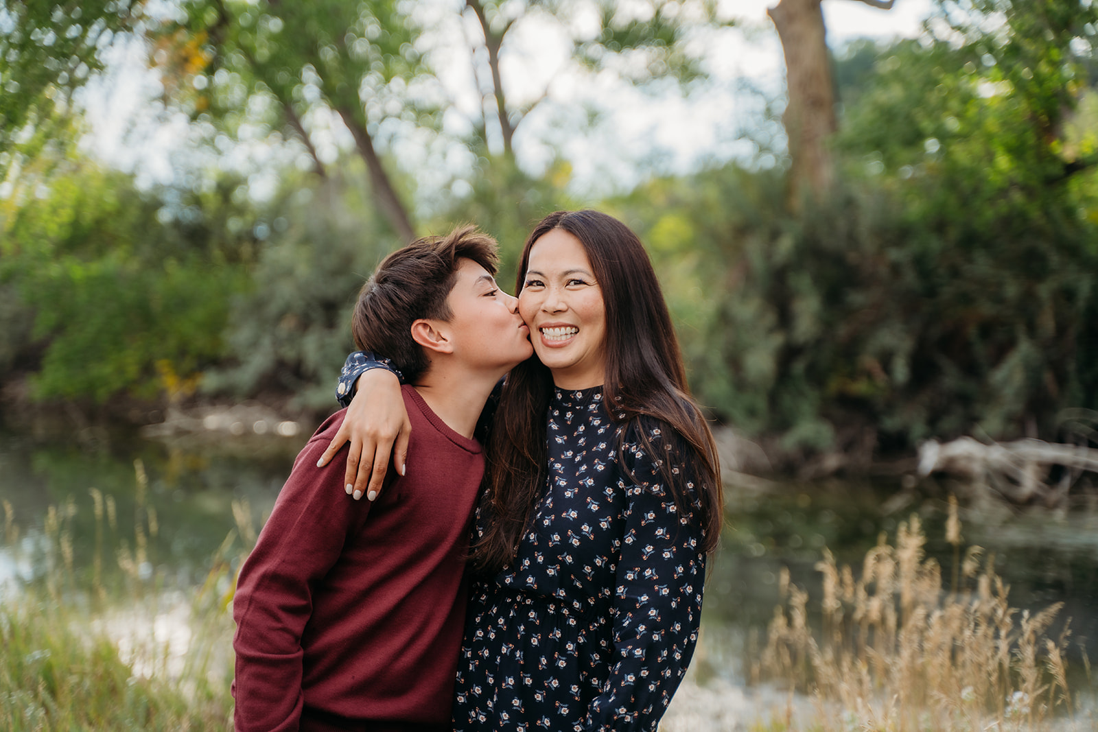 Mom beams as her son leans in for a kiss, a sweet candid from their family outdoor pictures.