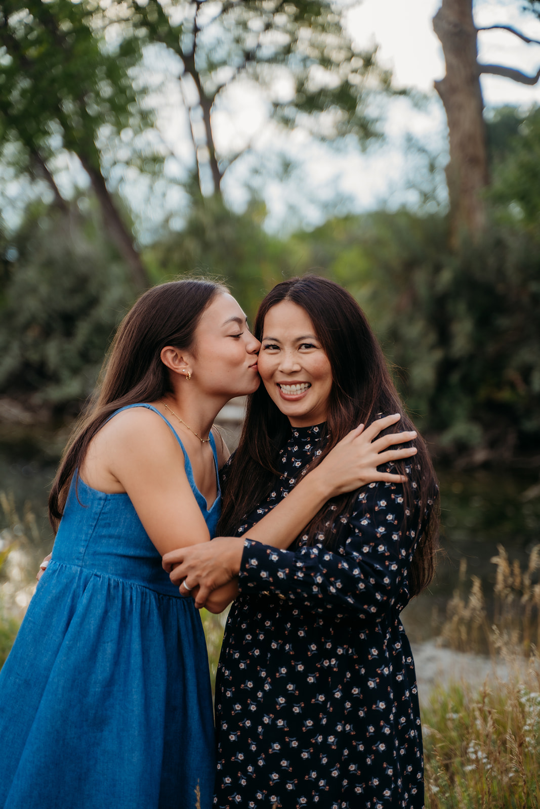 A kiss on the cheek between mother and daughter, surrounded by tall grasses and late-summer light.