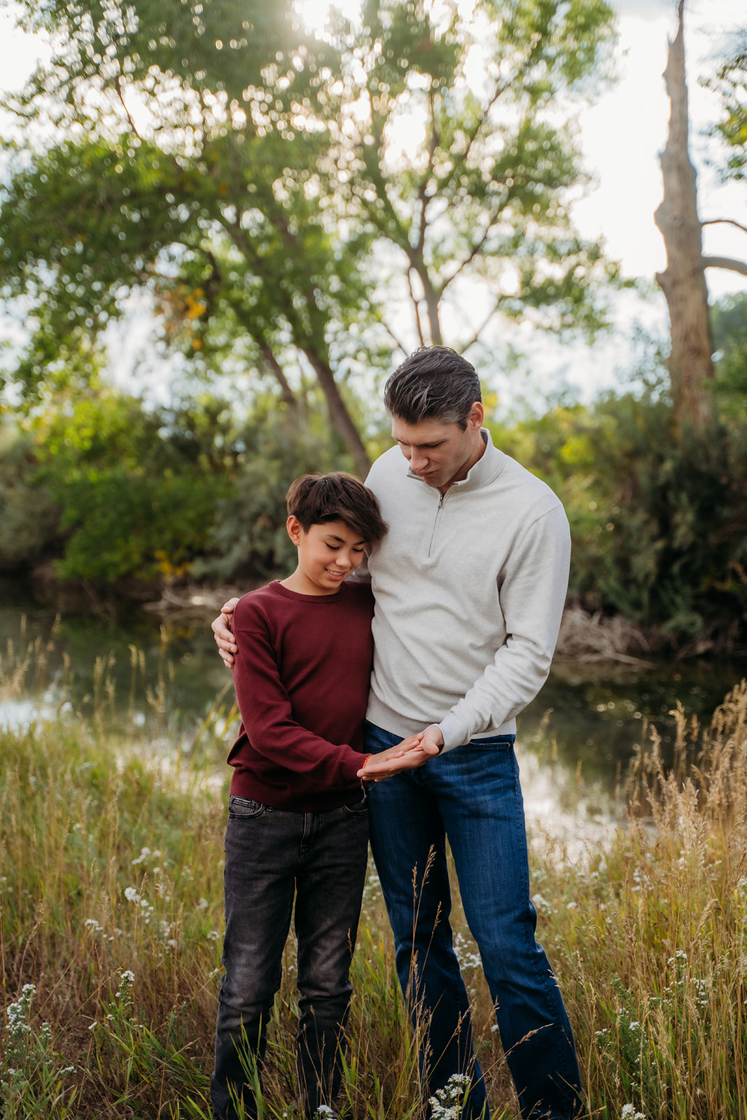 A dad and son share a tender moment by the water’s edge, heads bowed close in quiet conversation.
