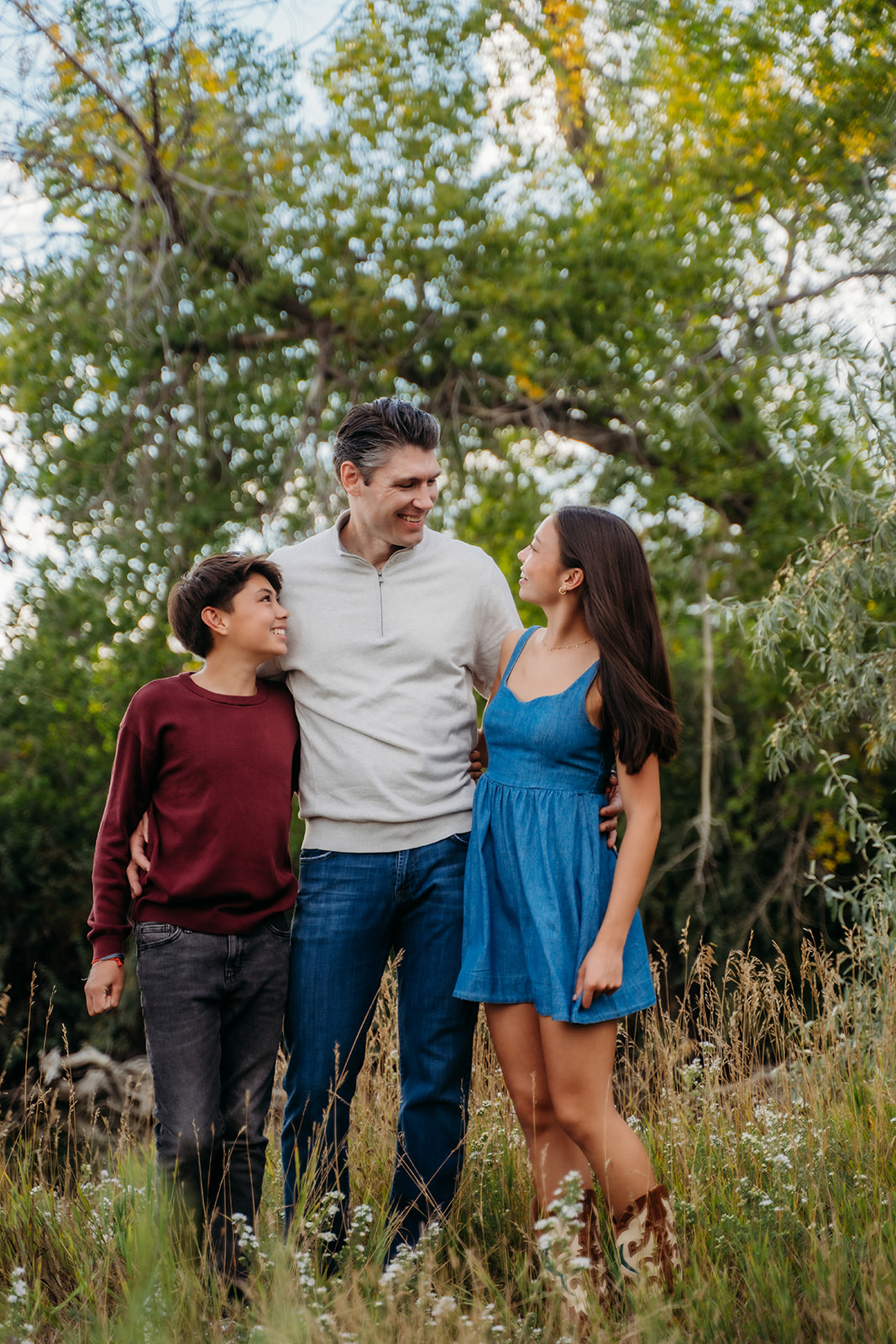 Dad shares a warm moment with his two kids, arms around their shoulders, all smiling beneath tall trees.