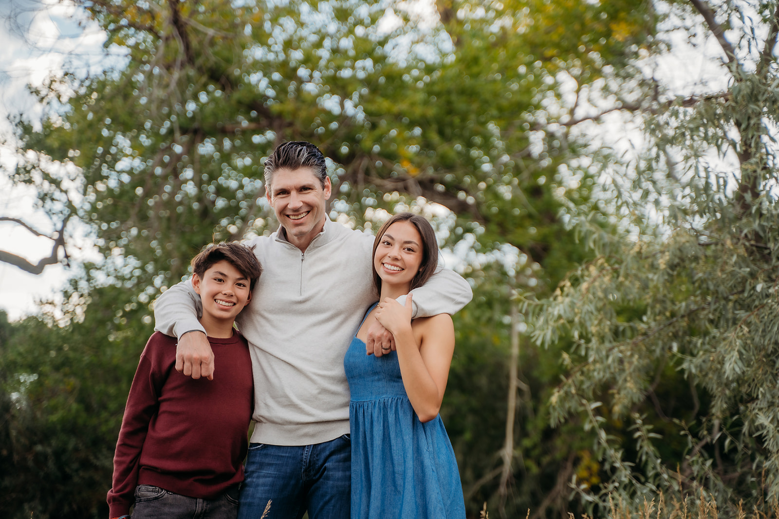 A proud dad wraps his arms around his two kids, the trio smiling brightly during their outdoor family session.