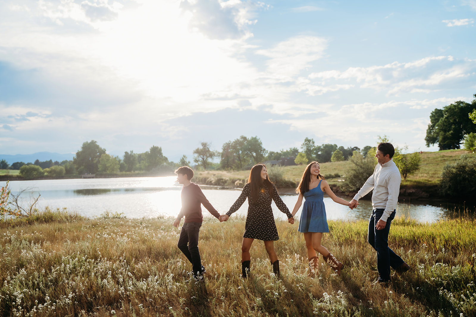 The family holds hands and walks through a sun-drenched meadow beside the lake, laughing together in the golden light.