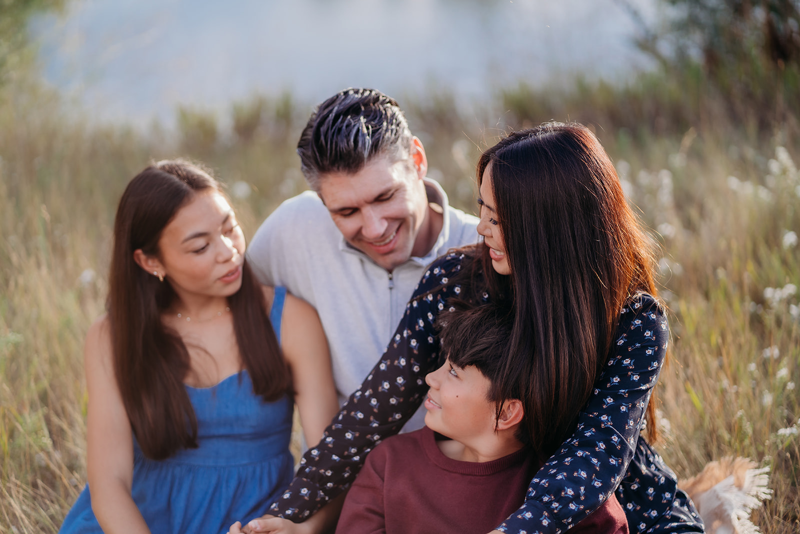 Sitting close in the tall grass, this family shares a quiet lakeside moment filled with smiles and sunshine.