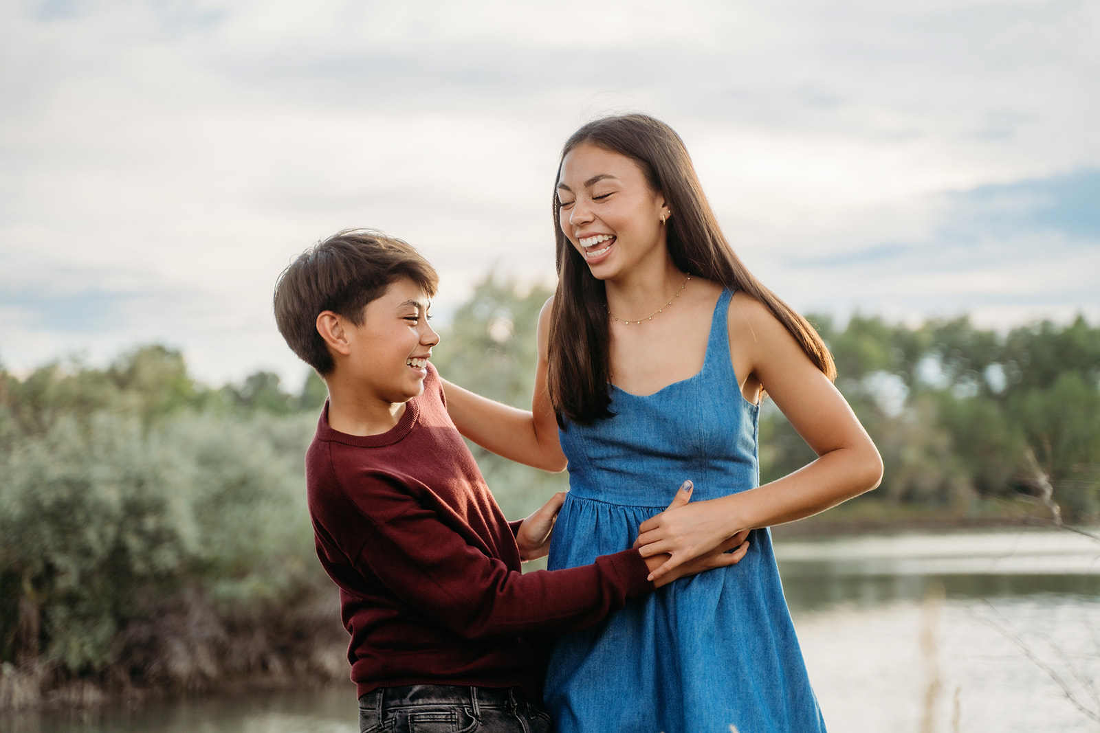 Sister and brother laugh together in front of a lake, their playful energy shining through in these sweet family outdoor pictures.