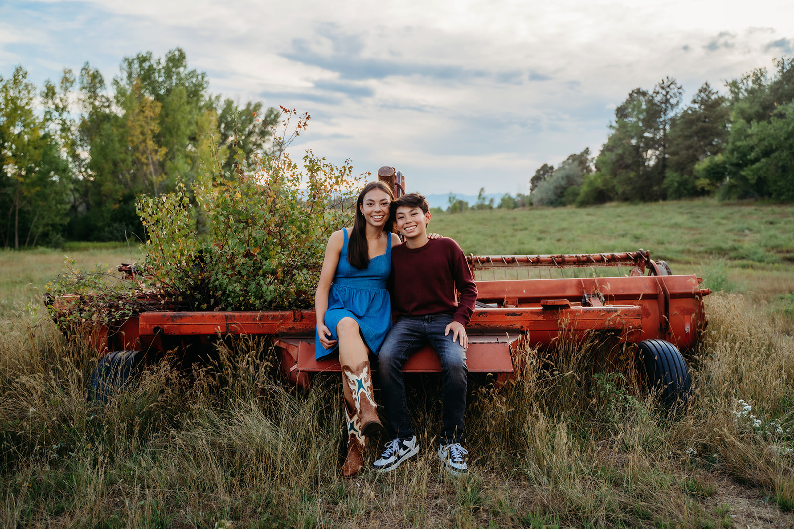 Seated on an old wagon in the middle of a wide-open field, the siblings smile with a mix of joy and comfort.