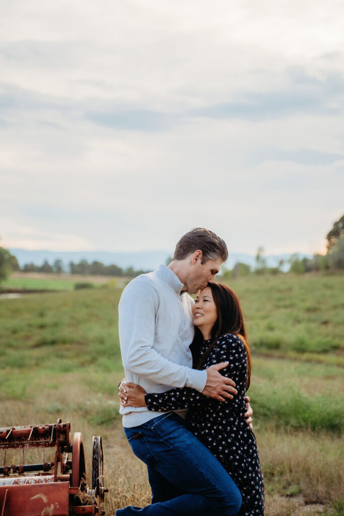 The couple embraces softly in the field, framed by mountains in the distance and warm golden hour skies.