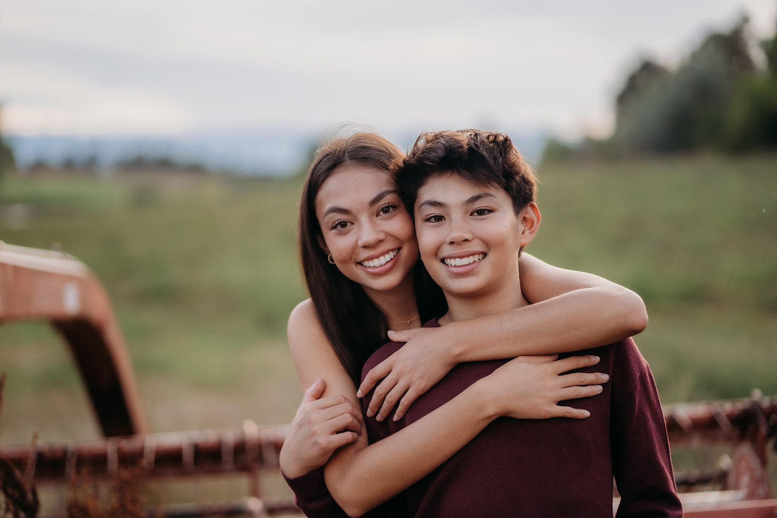 Big sister wraps her arms around her little brother as they smile wide, standing in front of a rustic wooden fence.