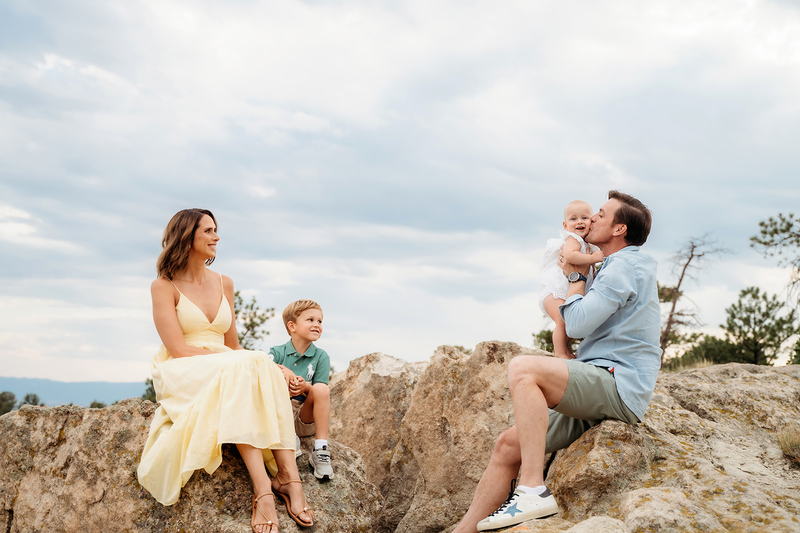 A family of four sitting on a rocky outcrop under a cloudy Colorado sky, sharing a relaxed and joyful moment together.