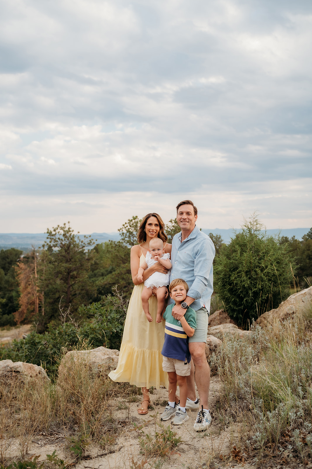 A smiling family of four posing together on a scenic overlook with trees and sky in the background—proof that relaxed vibes are key in family photo tips.