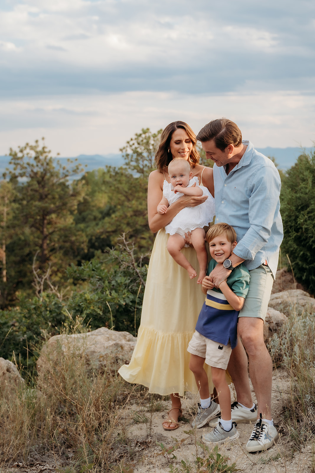 Parents holding their kids and laughing during a mountaintop photo session, framed by trees and golden hour light.