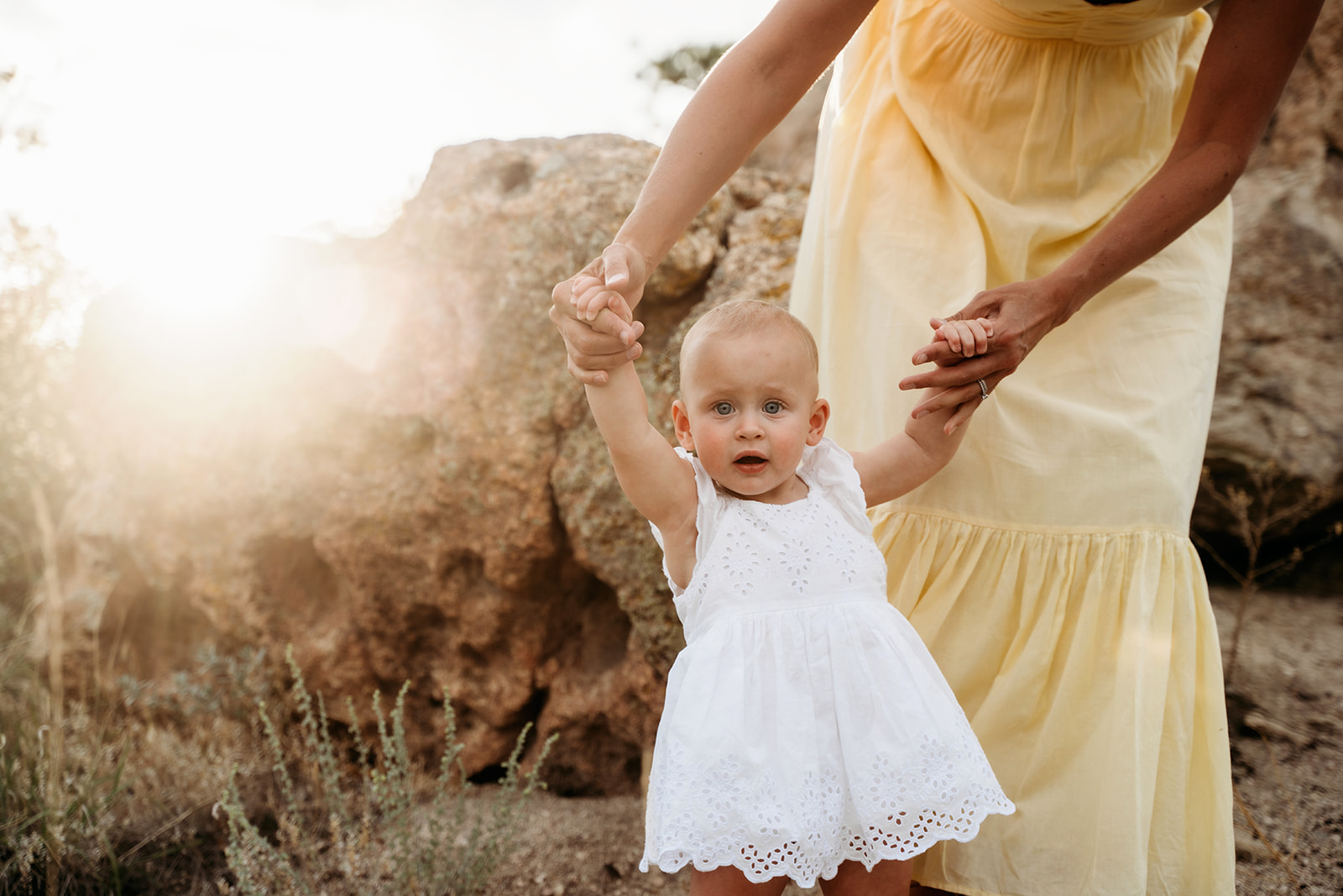 Toddler girl in a white eyelet dress holding her mother’s hands while walking near rocky terrain in natural light.