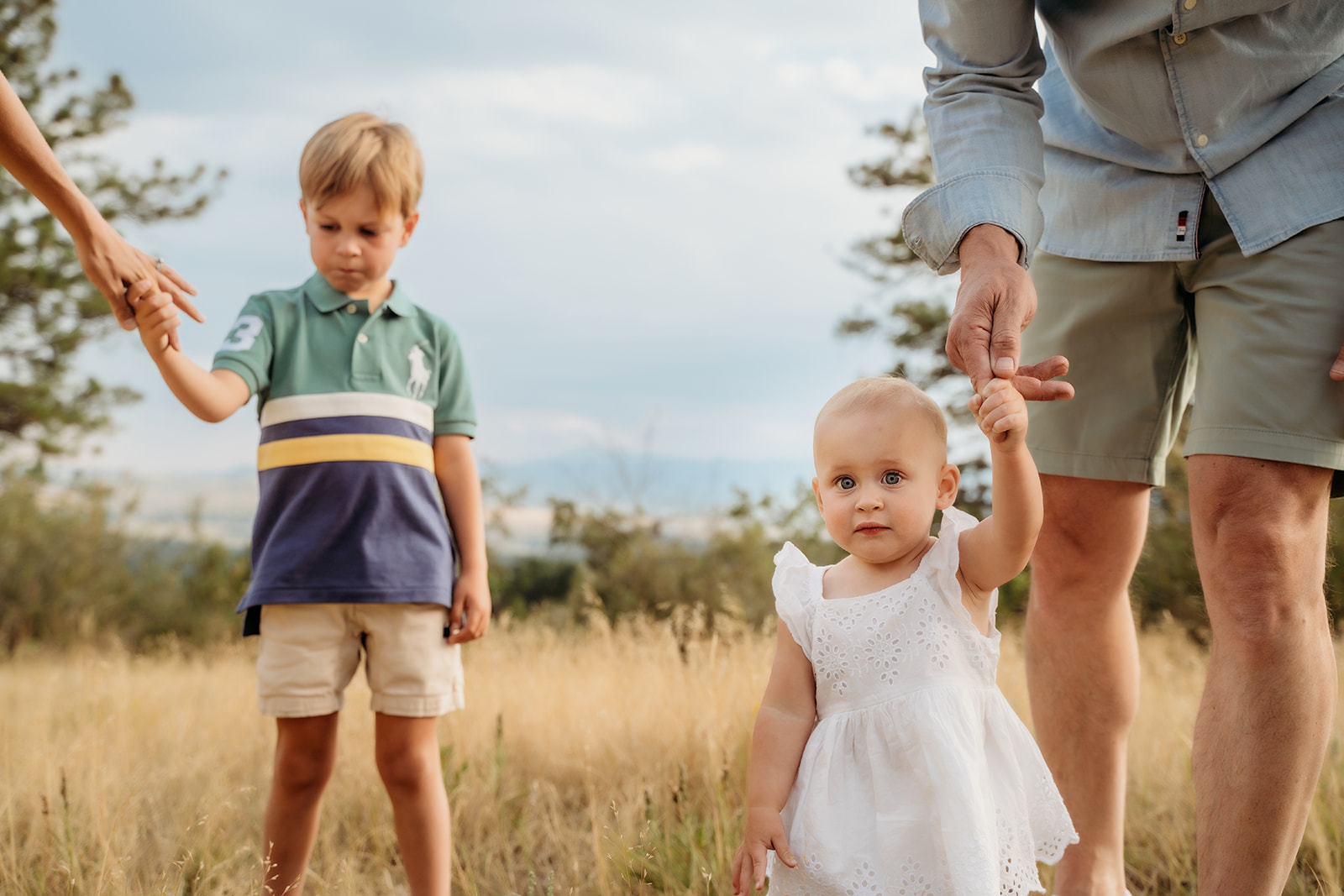 Baby girl holding hands with dad and walking confidently in a sunlit field, while her brother reaches for mom’s hand nearby.