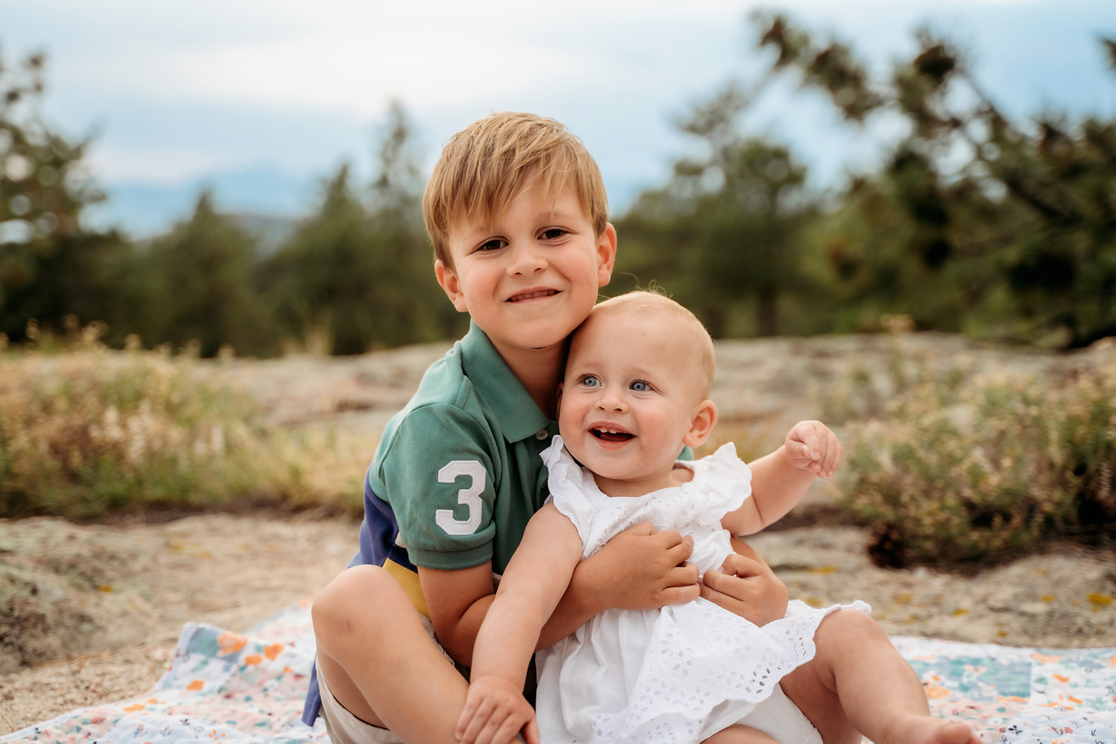 Big brother sitting on a quilt and holding his baby sister, both smiling sweetly during a candid moment outdoors.