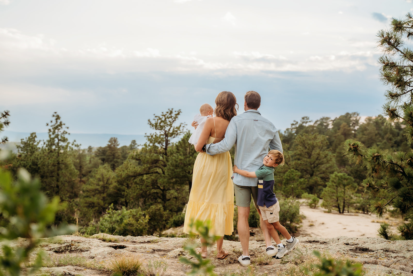 A family stands arm in arm overlooking a forested valley—one of the simplest family photo tips is to capture the view from behind for natural emotion.