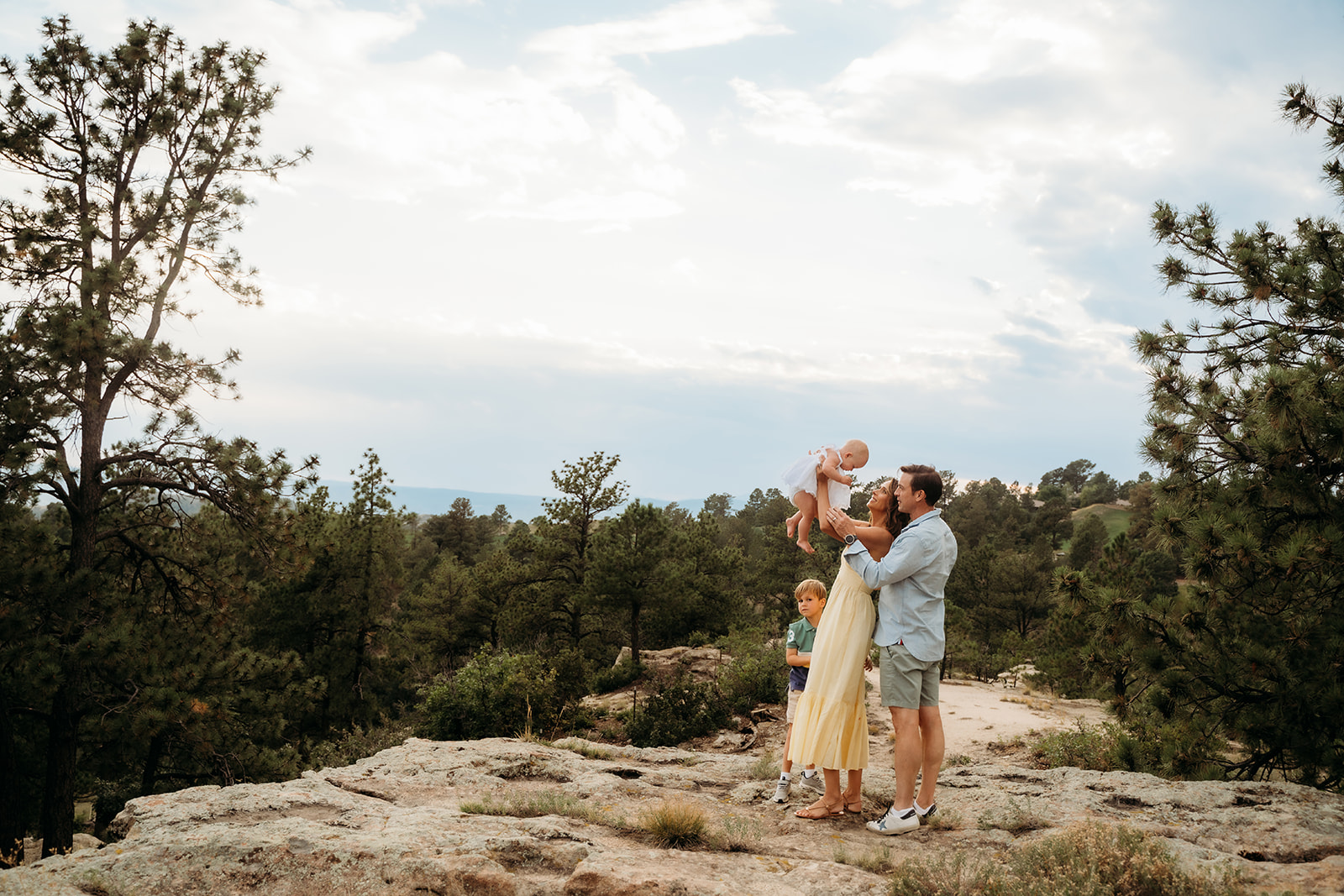 Dad lifts baby daughter toward the sky as mom and son look on, with pine trees and cloudy skies surrounding them—spontaneity is key in family photo tips.