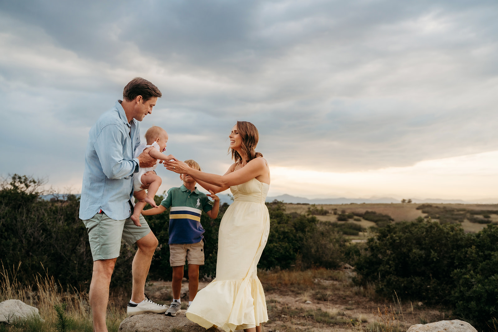 Dad handing baby daughter to mom during a golden hour session in the Colorado foothills, with big brother smiling in the background—natural interaction is one of the most important family photo tips.