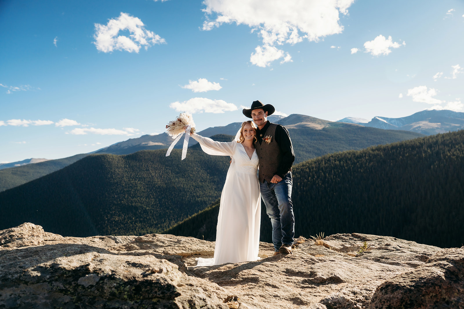 Bride and groom stand arm in arm on a mountaintop, bride raising her bouquet with a joyful grin—celebrating love, adventure, and learning how to write vows that reflect it all.