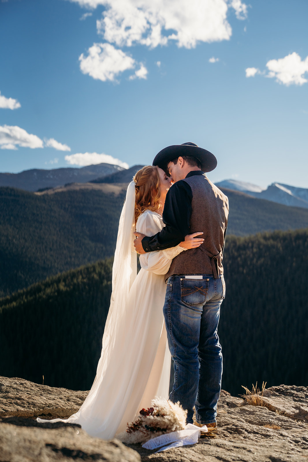 Couple shares a kiss on a rocky mountain ledge, bouquet and vow books resting at their feet.