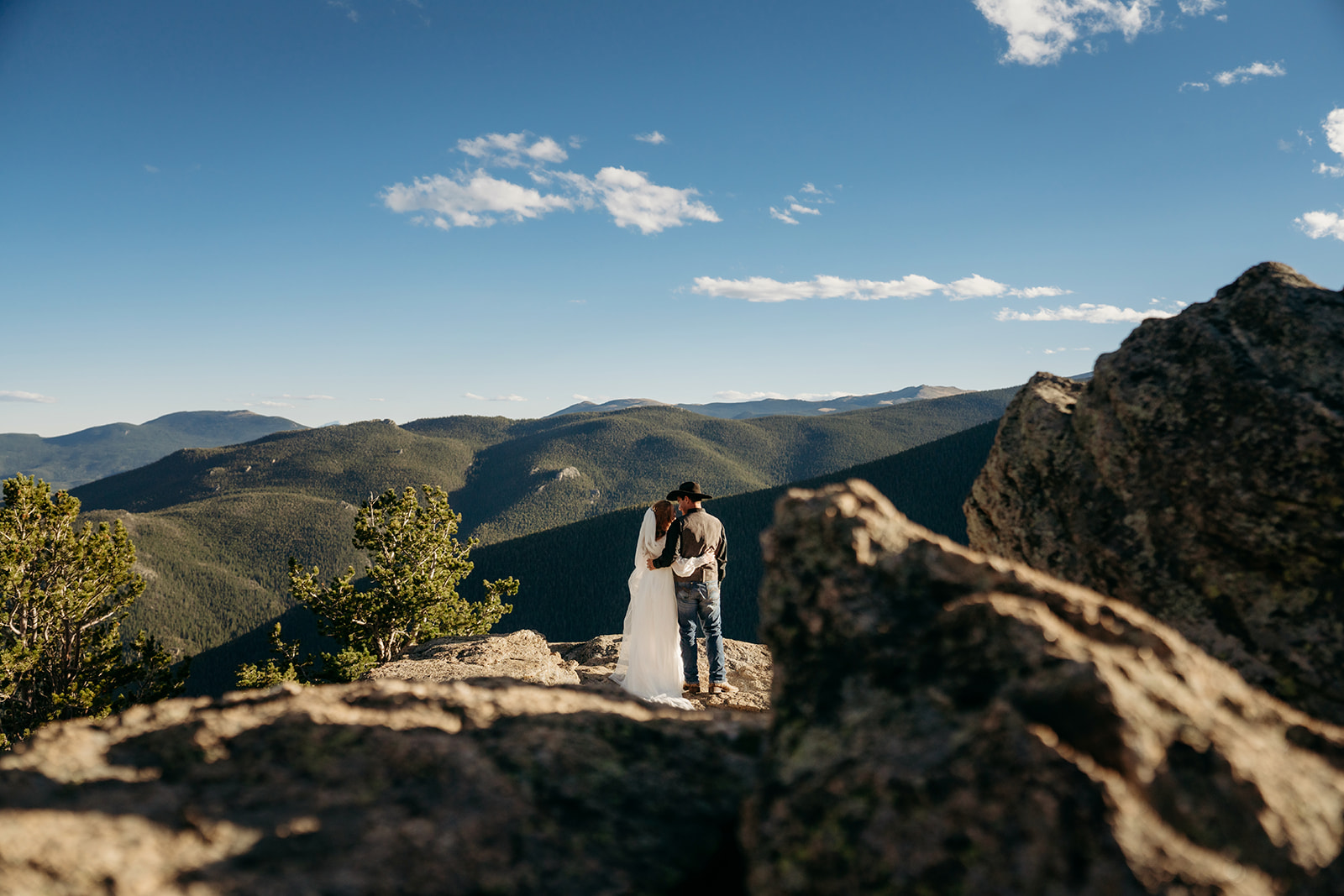 Wide-angle view of a bride and groom standing on a mountain ledge, silhouetted against the expansive Colorado landscape.