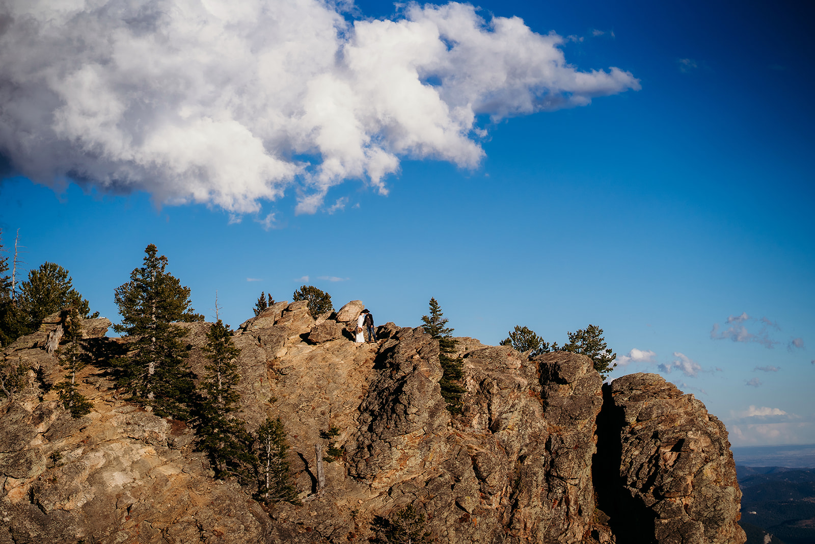 Wide shot of couple standing atop dramatic Colorado rock formations, surrounded by pine trees and blue skies.