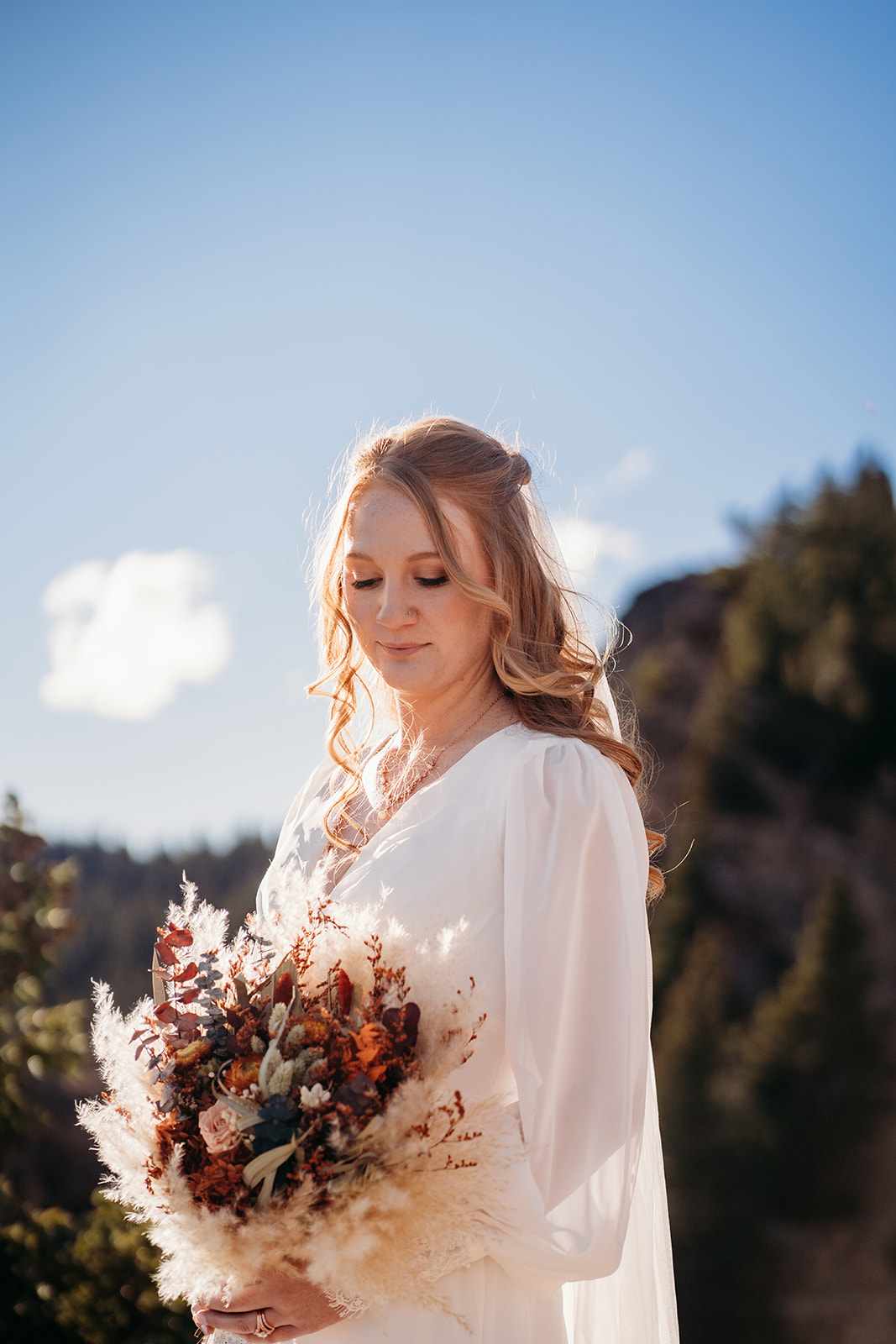 Bride stands softly lit by the afternoon sun, holding a bouquet of pampas grass and warm fall florals.