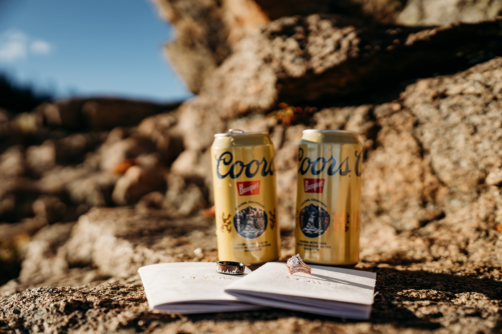 Close-up of vow books, wedding rings, and Coors Banquet beers on rock—how to write vows paired with Colorado style and personality.