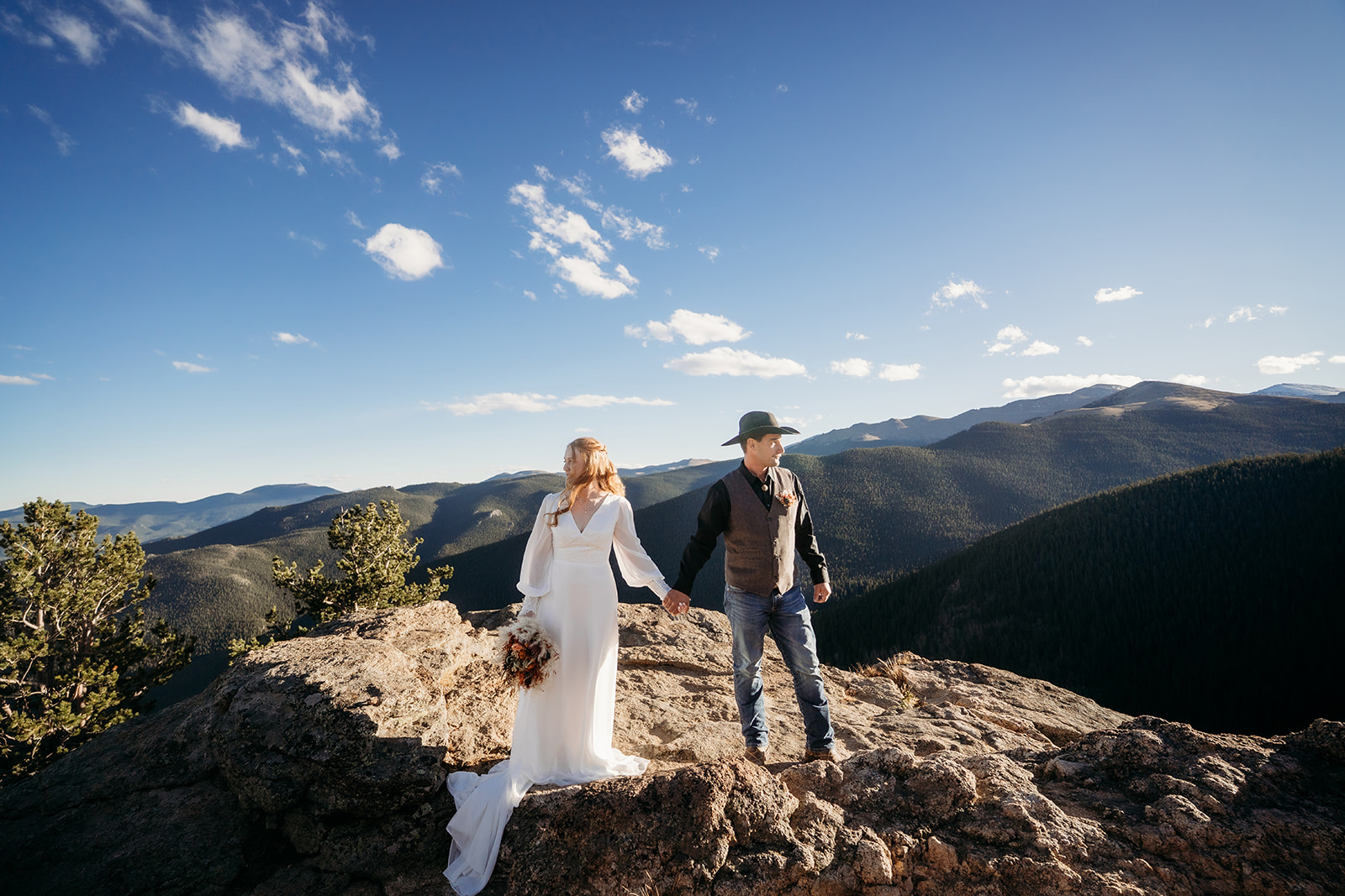 Bride and groom walk hand-in-hand across a rocky overlook, mountains stretching out endlessly behind them.