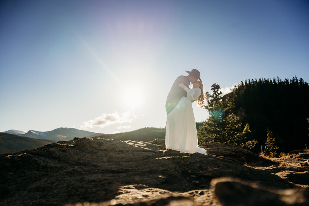 Groom dips bride for a kiss on a sunny mountaintop, surrounded by evergreen trees and Colorado peaks.