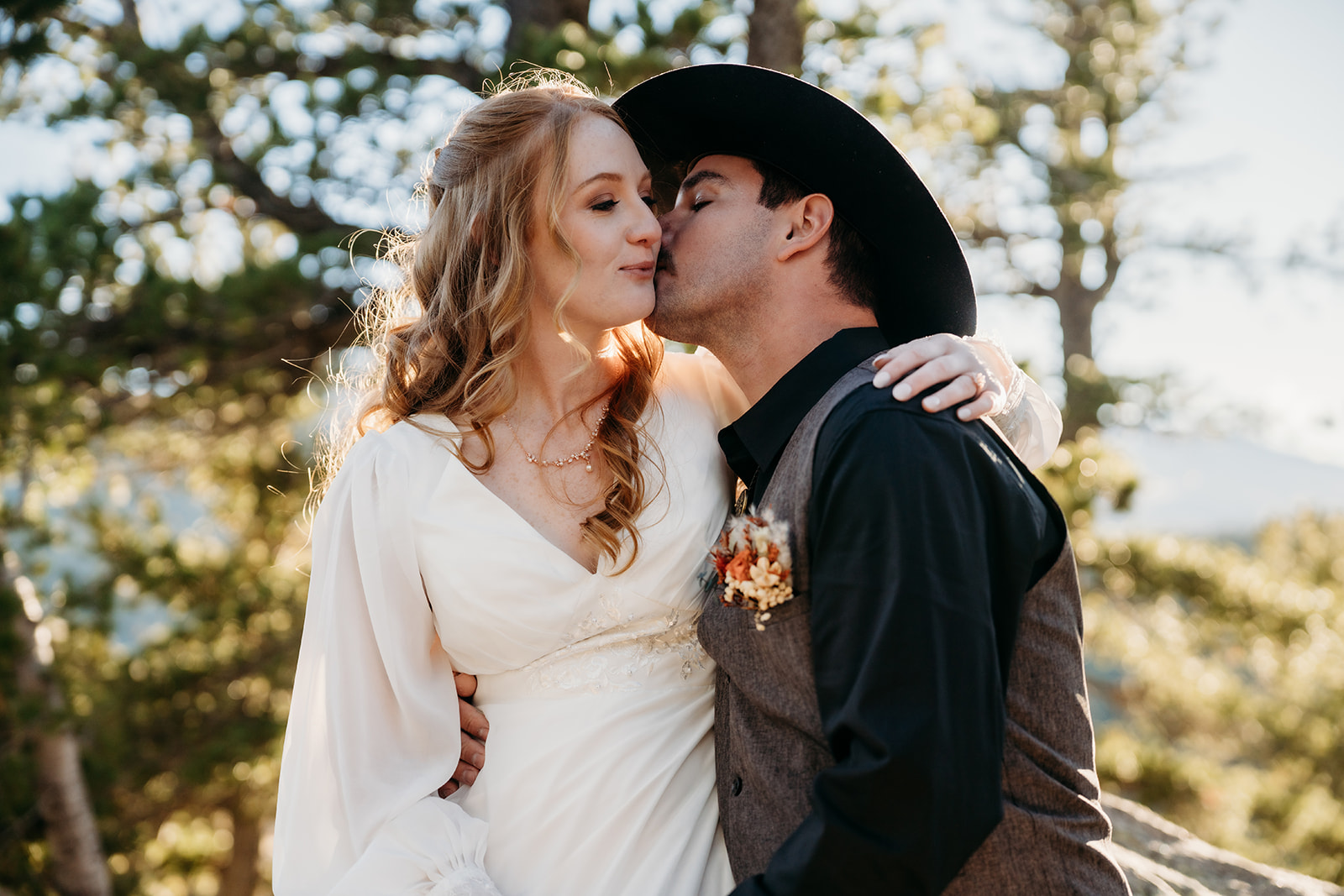 Bride smiles as the groom kisses her cheek under pine trees, a sweet moment following their elopement ceremony.