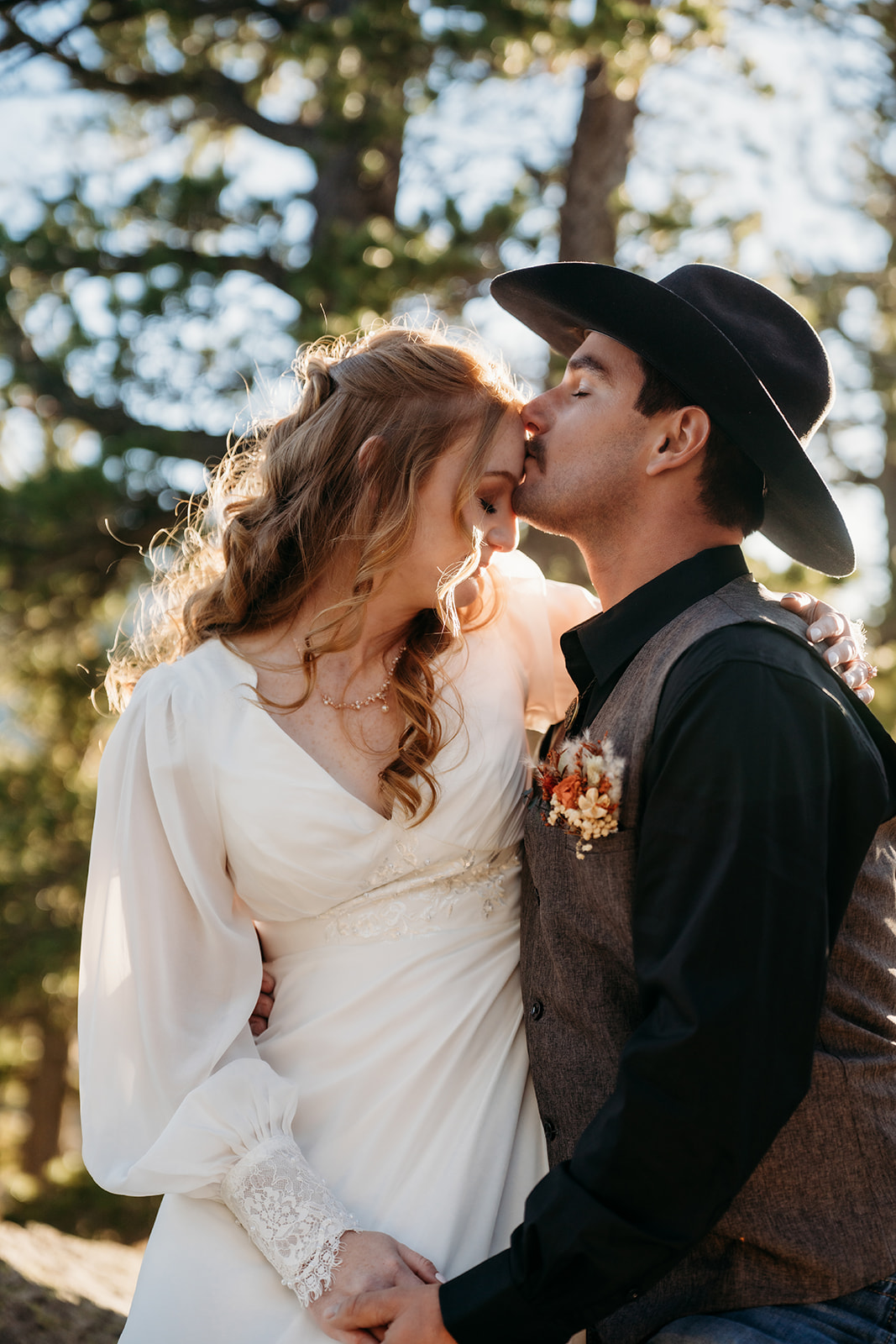 Groom kisses bride’s forehead in soft afternoon light, surrounded by trees during their intimate elopement.
