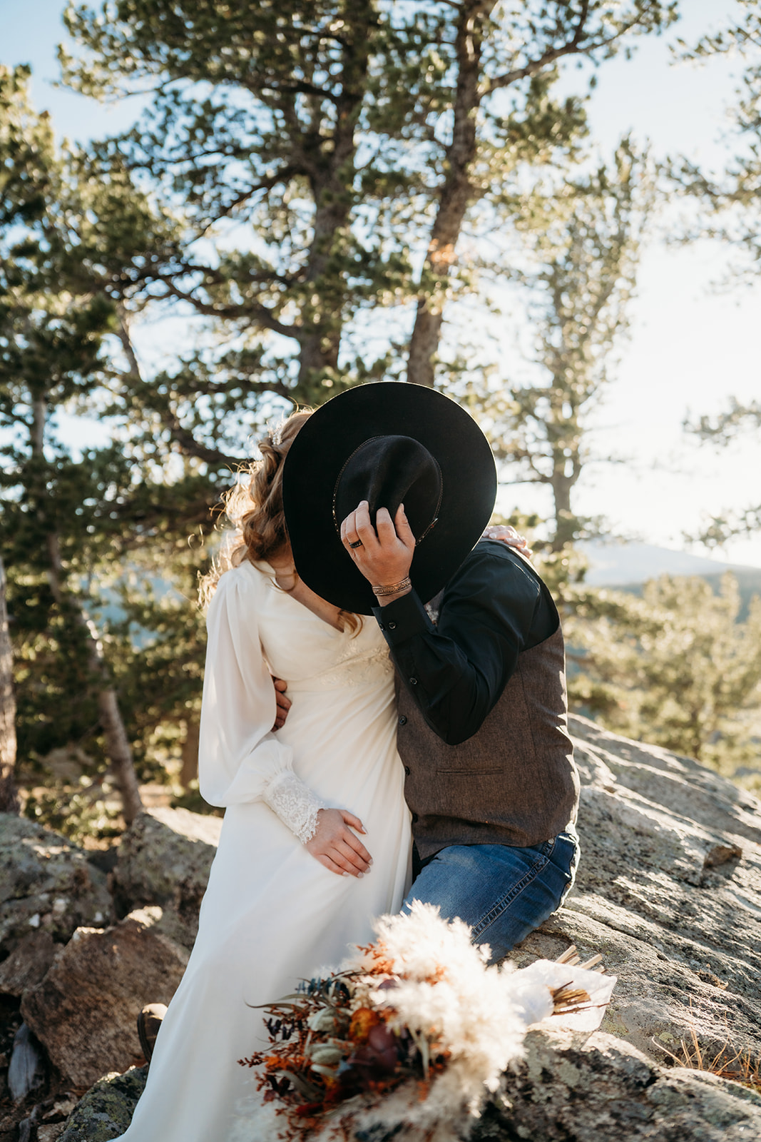 Bride and groom share a playful kiss behind his cowboy hat, bouquet of dried florals resting on nearby rocks.