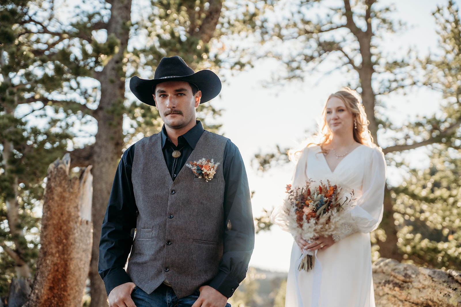 Groom stands in the forest with a serious expression as his bride smiles behind him, holding a bouquet of dried florals—setting the scene for how to write vows that are both playful and heartfelt.