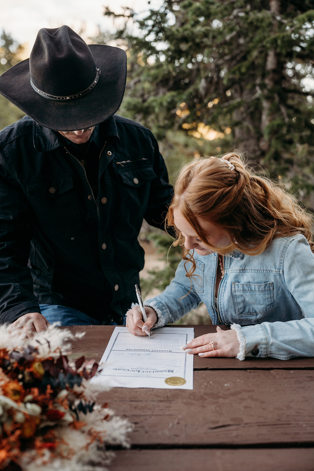 Bride signs the marriage license at a rustic wooden table, surrounded by fall florals after their heartfelt vows.