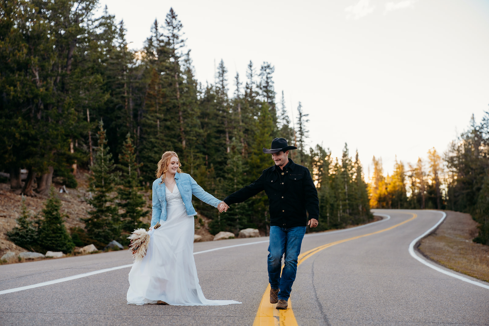 Bride and groom laugh and hold hands while walking down a forest road, radiating joy after their mountaintop wedding.