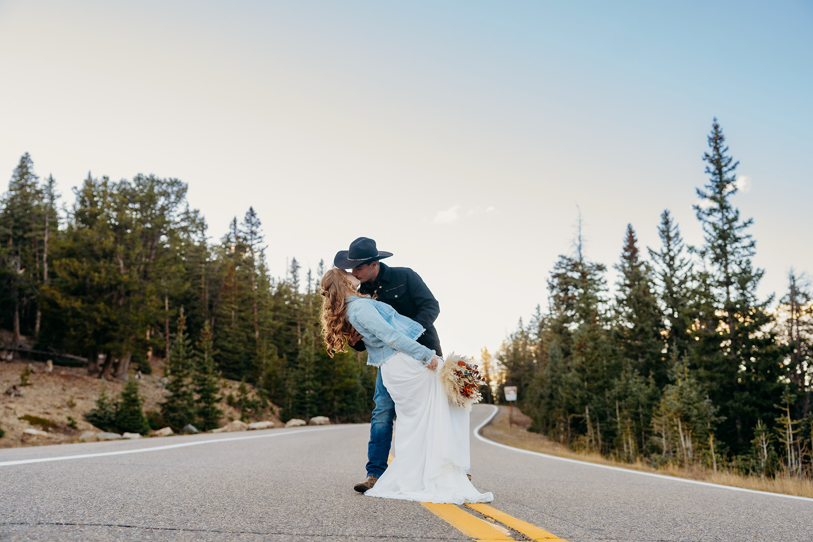 Groom dips bride for a kiss in the middle of a winding mountain road, her bouquet and denim jacket adding a laid-back touch.