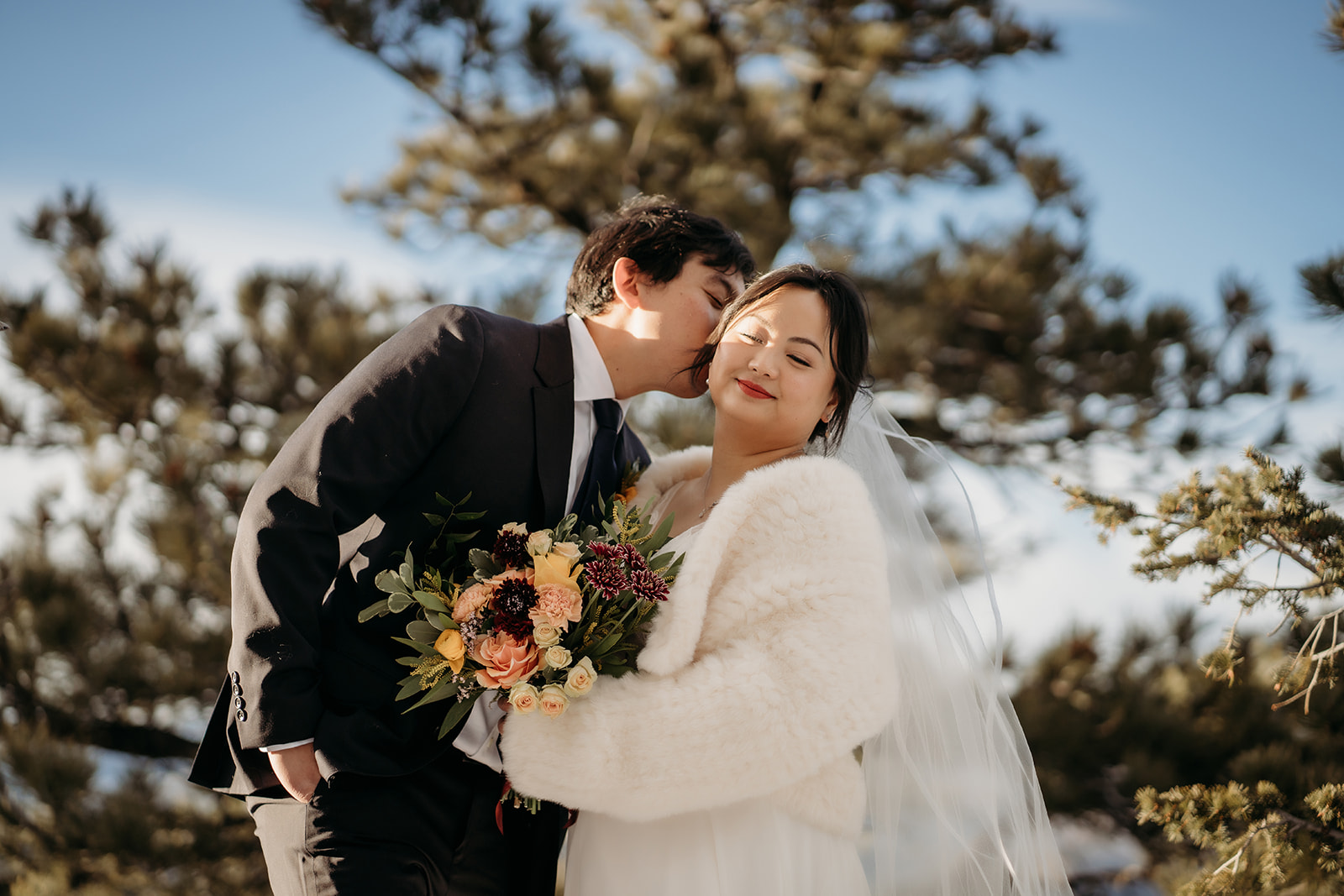 Groom kisses bride on the temple as she holds a seasonal bouquet and smiles in her faux fur wrap.