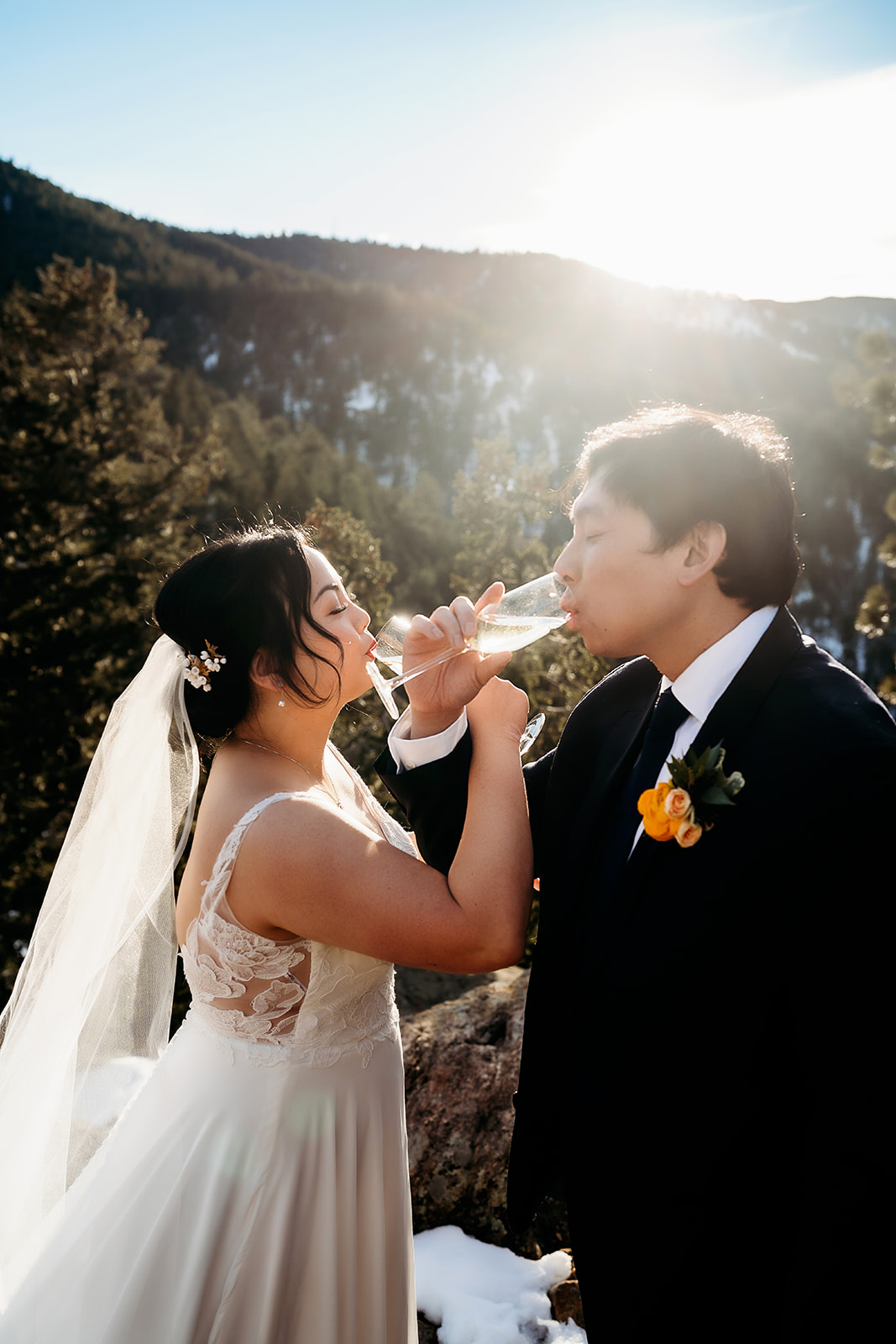 Newlyweds share a champagne toast in the mountains, sunlight glowing behind them.