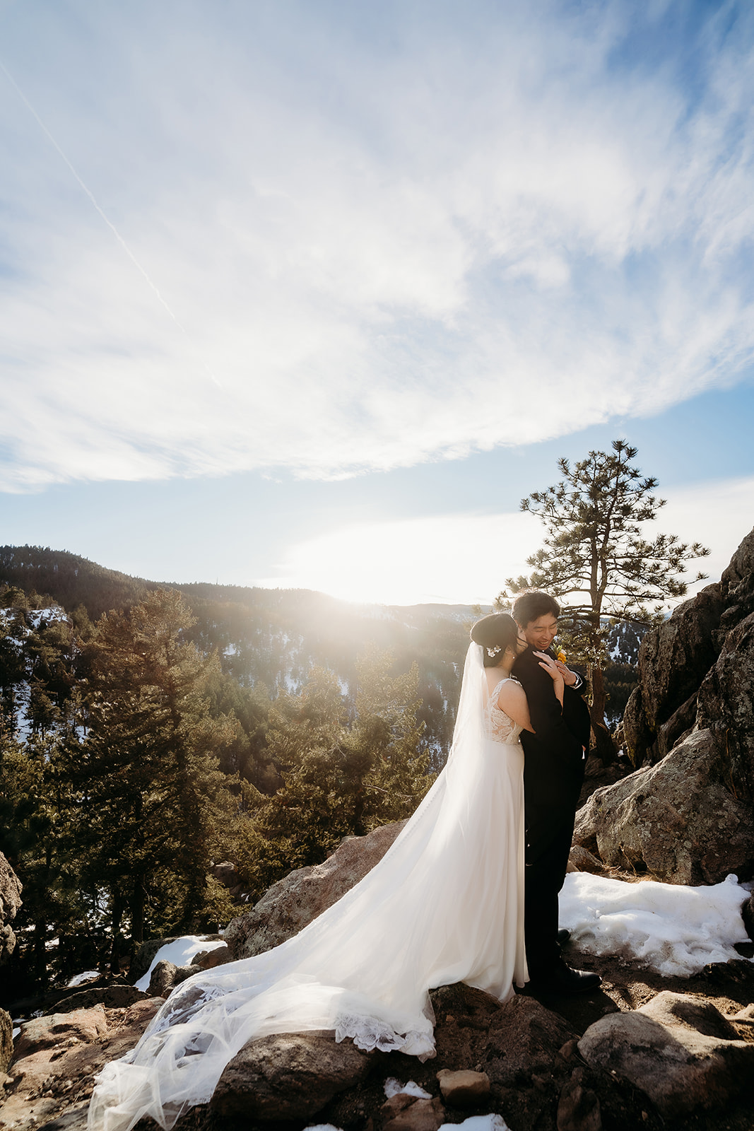 Couple stands arm-in-arm on a sunlit mountain ledge surrounded by snowy pine trees.