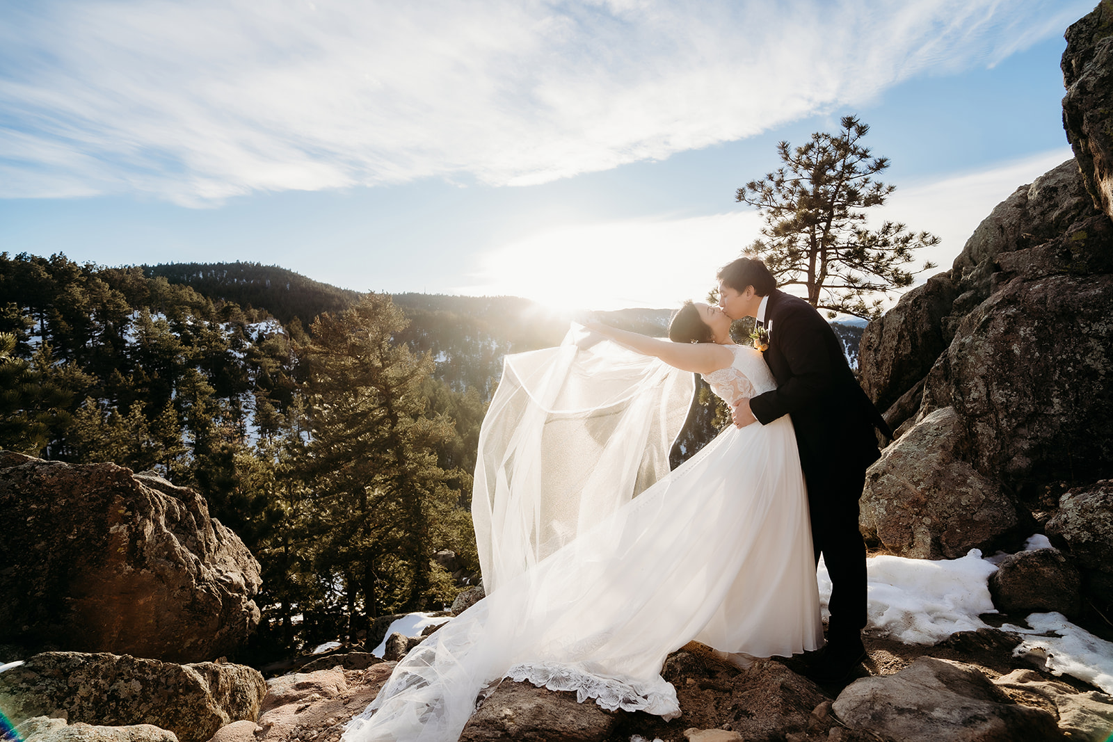 Romantic kiss on a rocky overlook as the bride’s veil flows in the wind during their Colorado winter wedding.