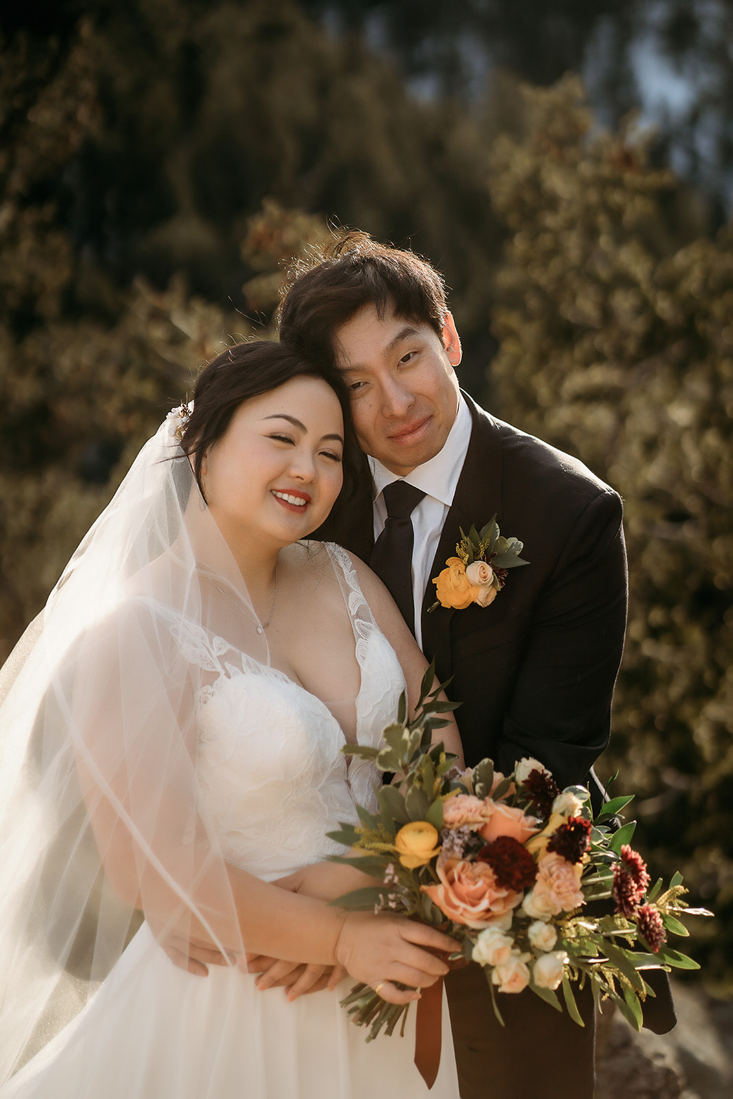 Bride and groom snuggle close, smiling with soft sunlight and forest backdrop.
