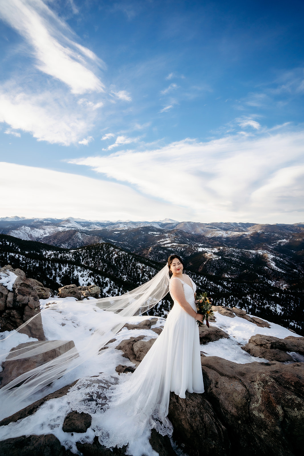 Bride smiles as her veil catches the wind on a mountaintop, framed by a snowy Colorado landscape.