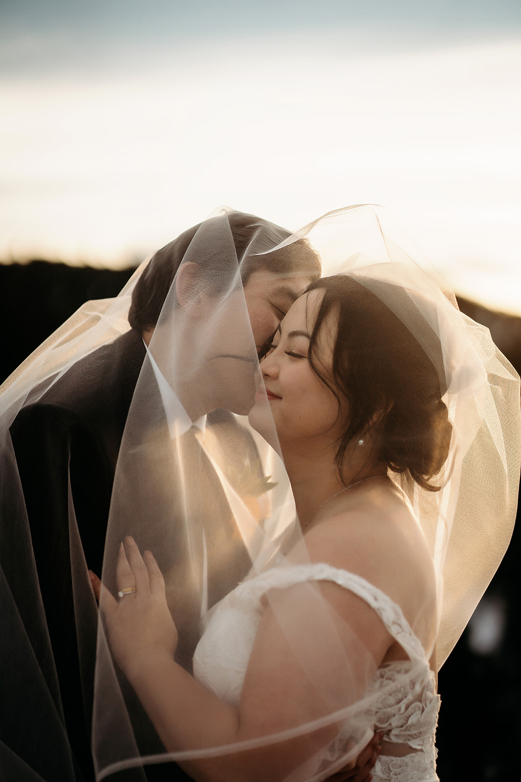 Romantic moment under the veil with soft sunset light illuminating the couple’s faces.