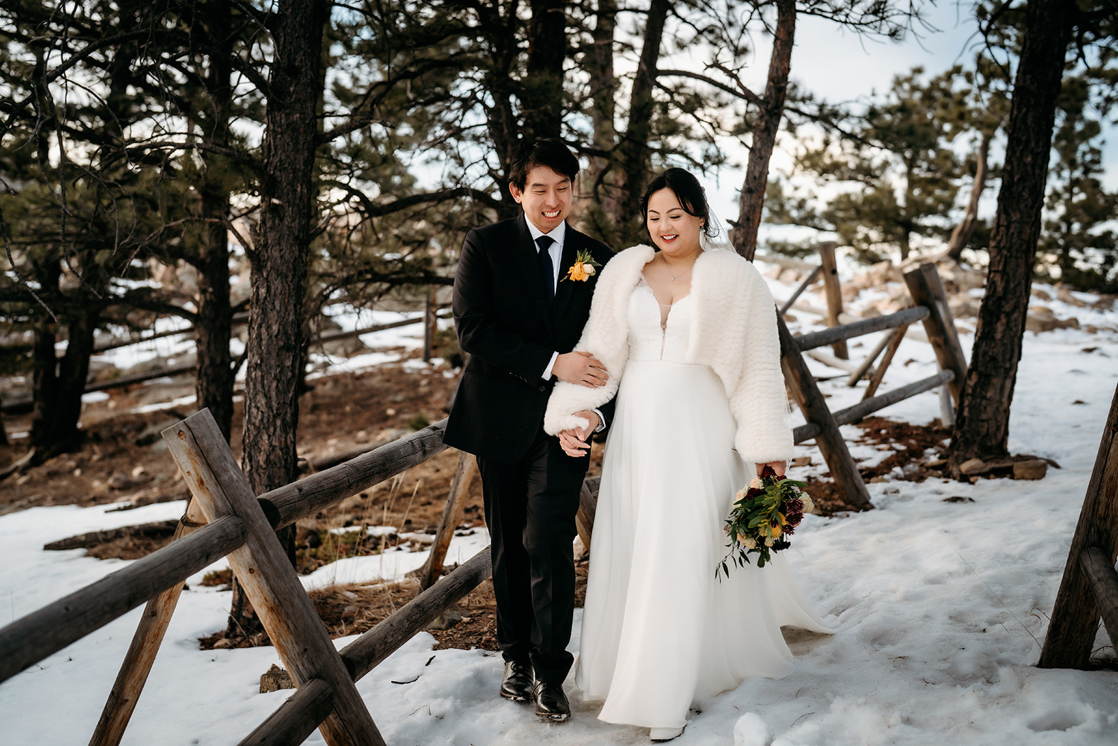 Newlyweds walking through a snowy forest trail, bundled up for their cozy winter wedding.