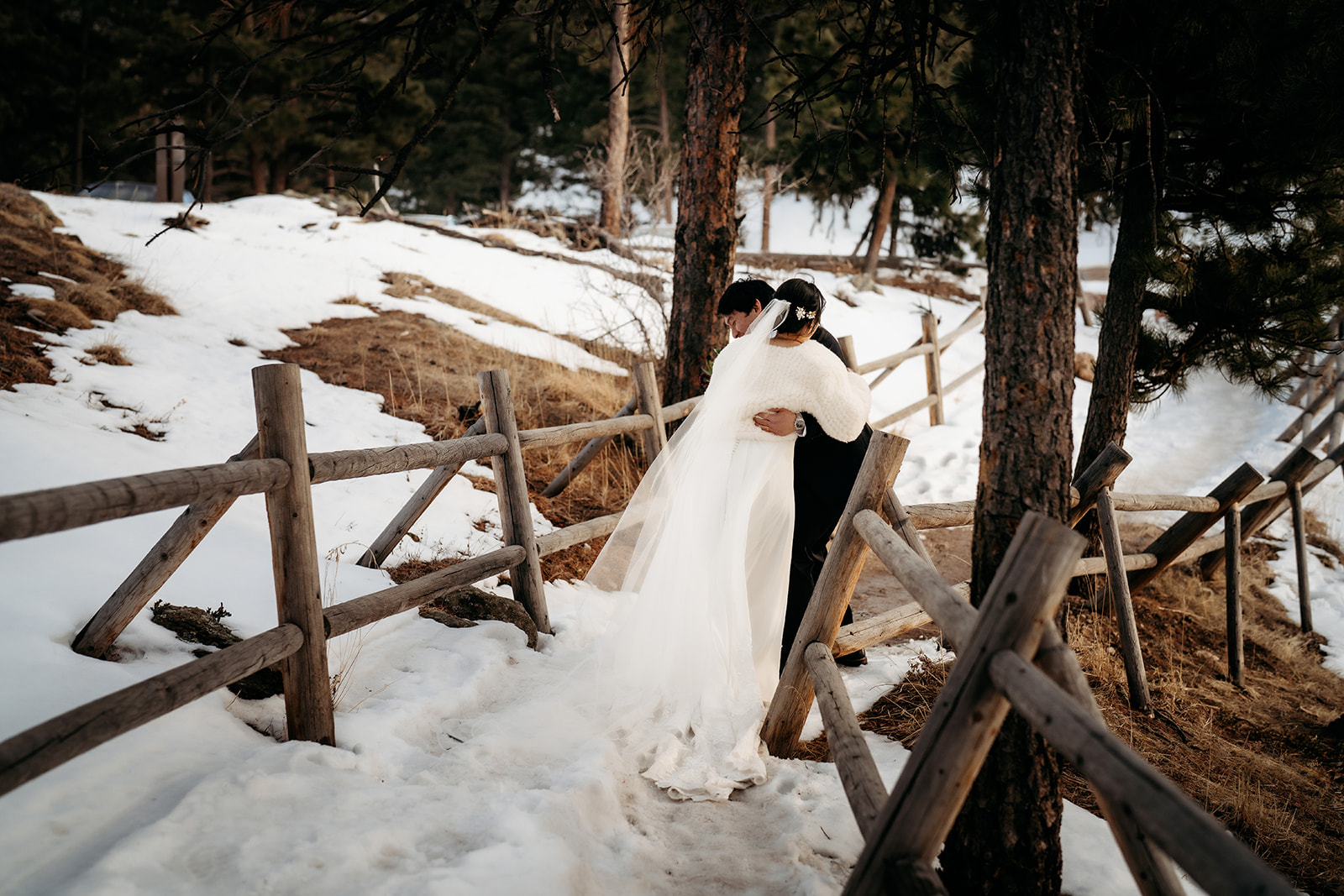 Couple walks a snow-lined path surrounded by wooden fencing and evergreens, bundled close together.