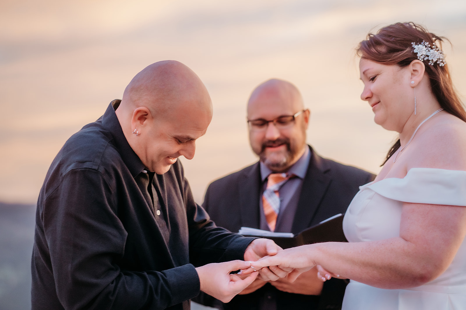 Groom placing a ring on the bride’s finger as their officiant smiles behind them—joyful, emotional, and beautifully captured at golden hour.