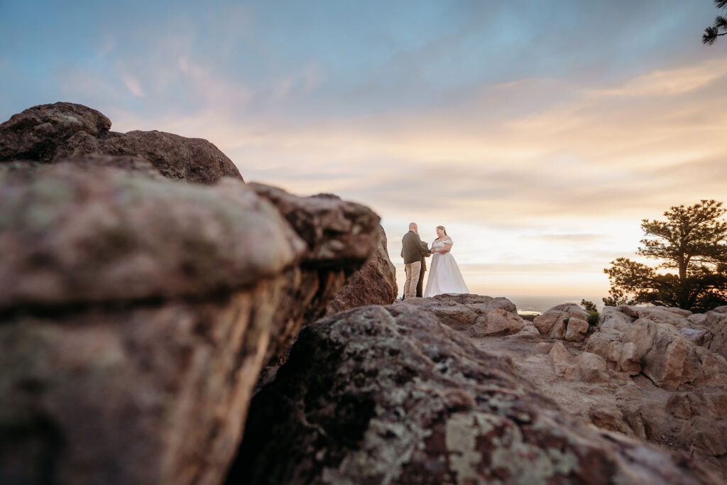 A wide-angle view of the couple’s ceremony from below the rocks, with dreamy pastel skies behind them.
