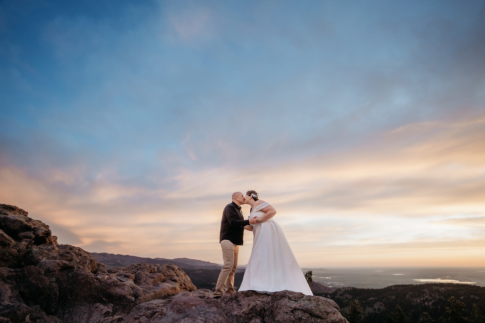 Bride and groom sharing a kiss atop a rocky mountain overlook at sunset, with dreamy skies captured by an elopement photographer in Colorado.