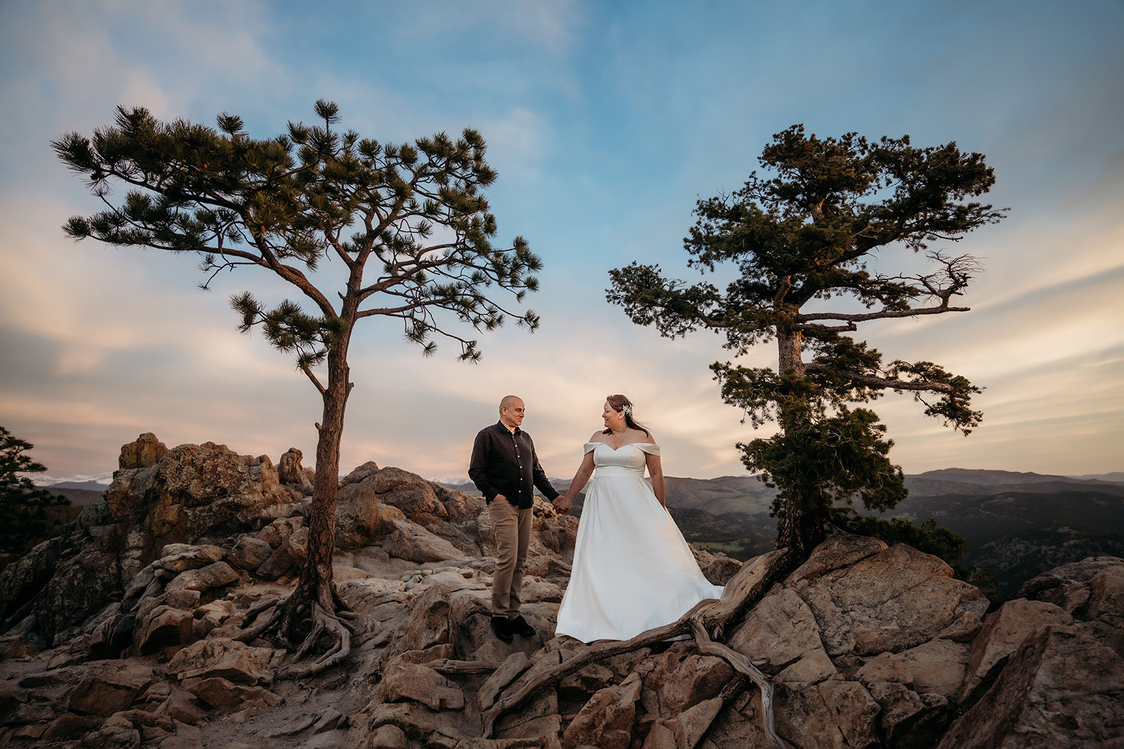Couple holds hands between two wind-swept trees, standing on rocky ground with mountains in the distance and the sky glowing at dusk.