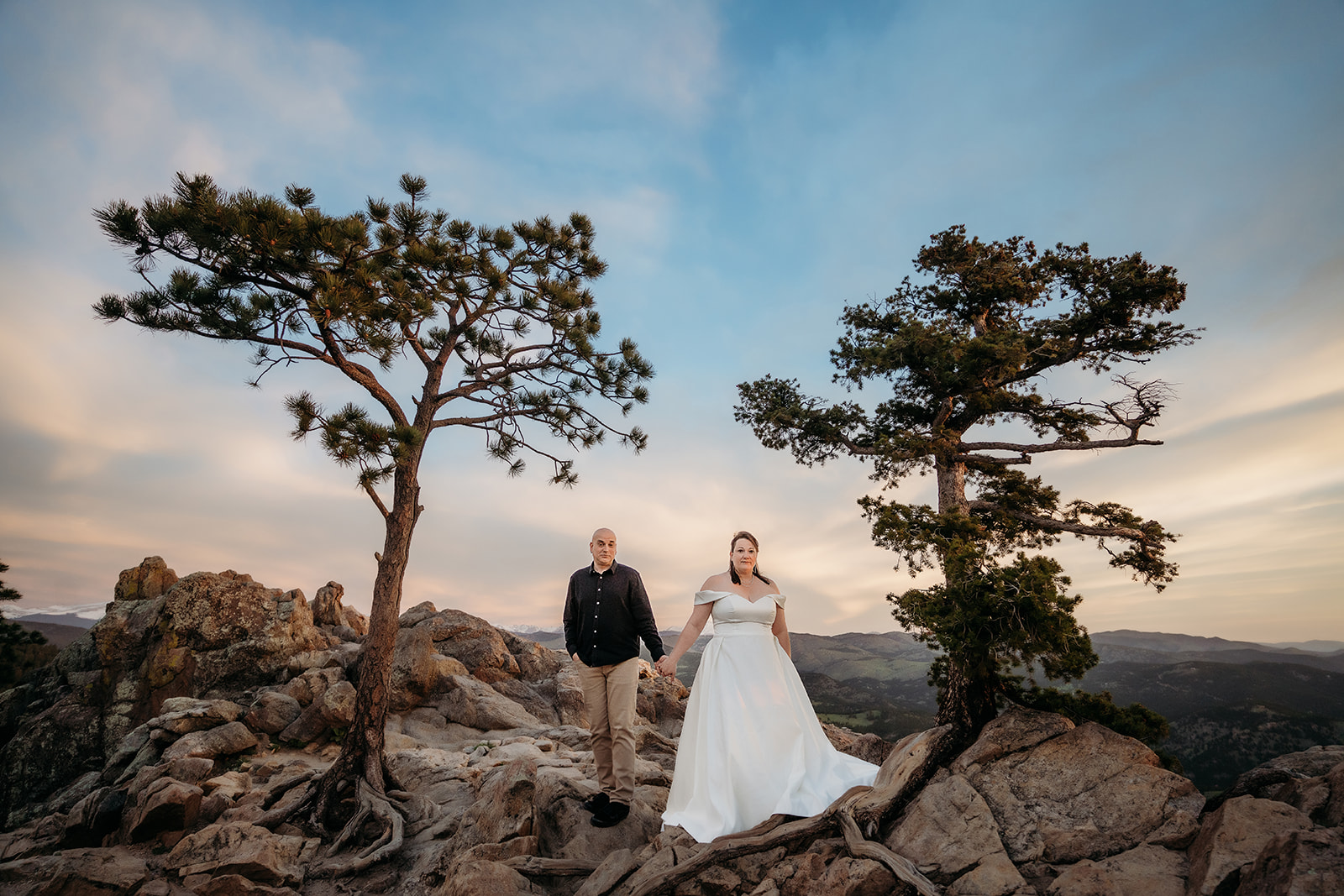 Couple stands hand in hand between two iconic trees on a rocky overlook, perfectly framed by their elopement photographer Colorado.
