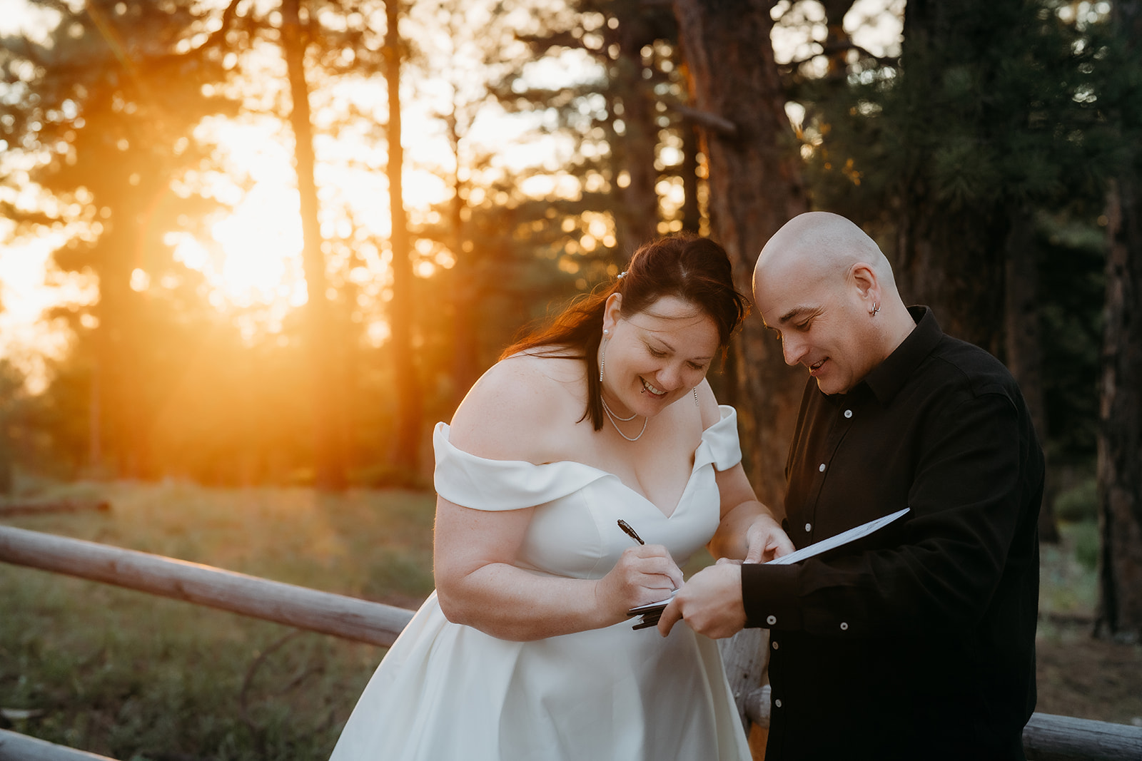 Bride signs the marriage license with a joyful laugh as the sun filters through the pine trees behind her.